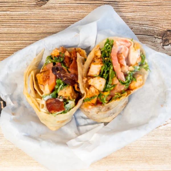 Two chicken wraps with bacon, lettuce, and sauce, served in white paper on a wooden table.