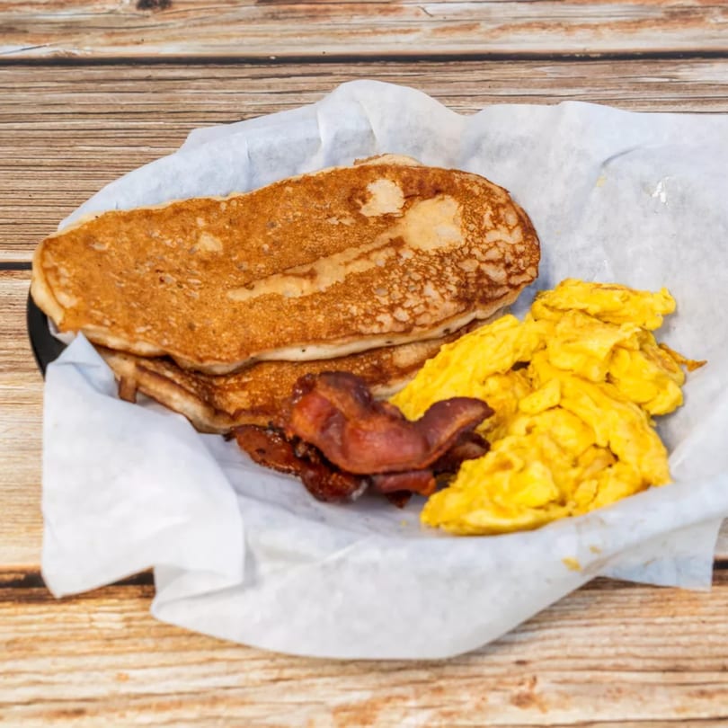 Pancakes, scrambled eggs, and bacon served in a white paper-lined basket on a wooden table.