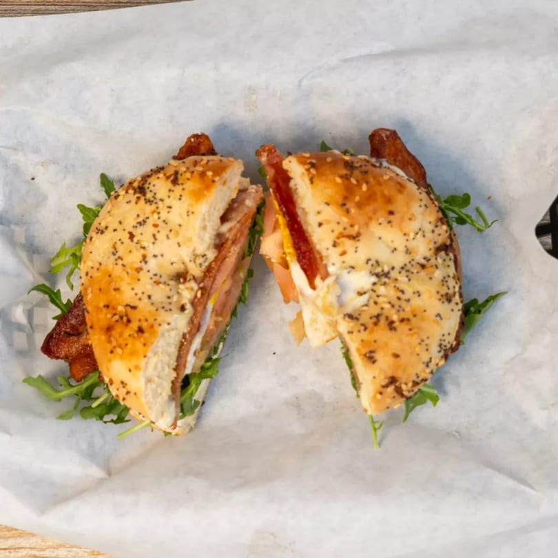 Everything bagel sandwich, cut in half, with bacon, egg, cheese, and arugula on parchment paper.