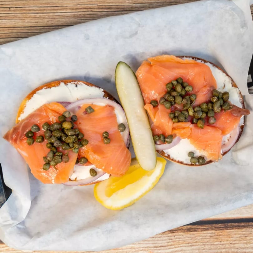 Two bagels with cream cheese, smoked salmon, capers, red onion, a pickle spear, and lemon.